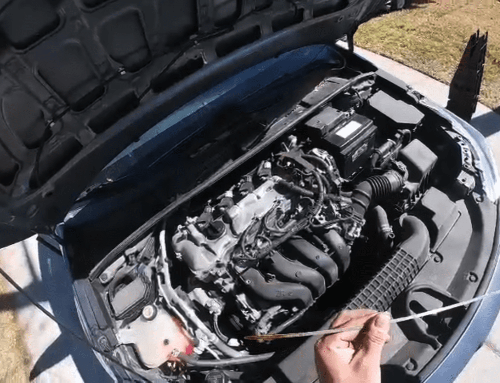 A close-up view of an open car engine with a person's hand pointing to or examining internal mechanical components under daylight.