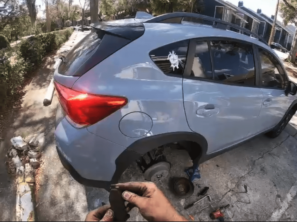 A person is working on removing or inspecting a tire from a light blue compact vehicle parked on a residential driveway surrounded by trees.
