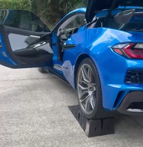 A bright blue sports car with its driver-side door open sits on a paved surface with trees visible in the background.