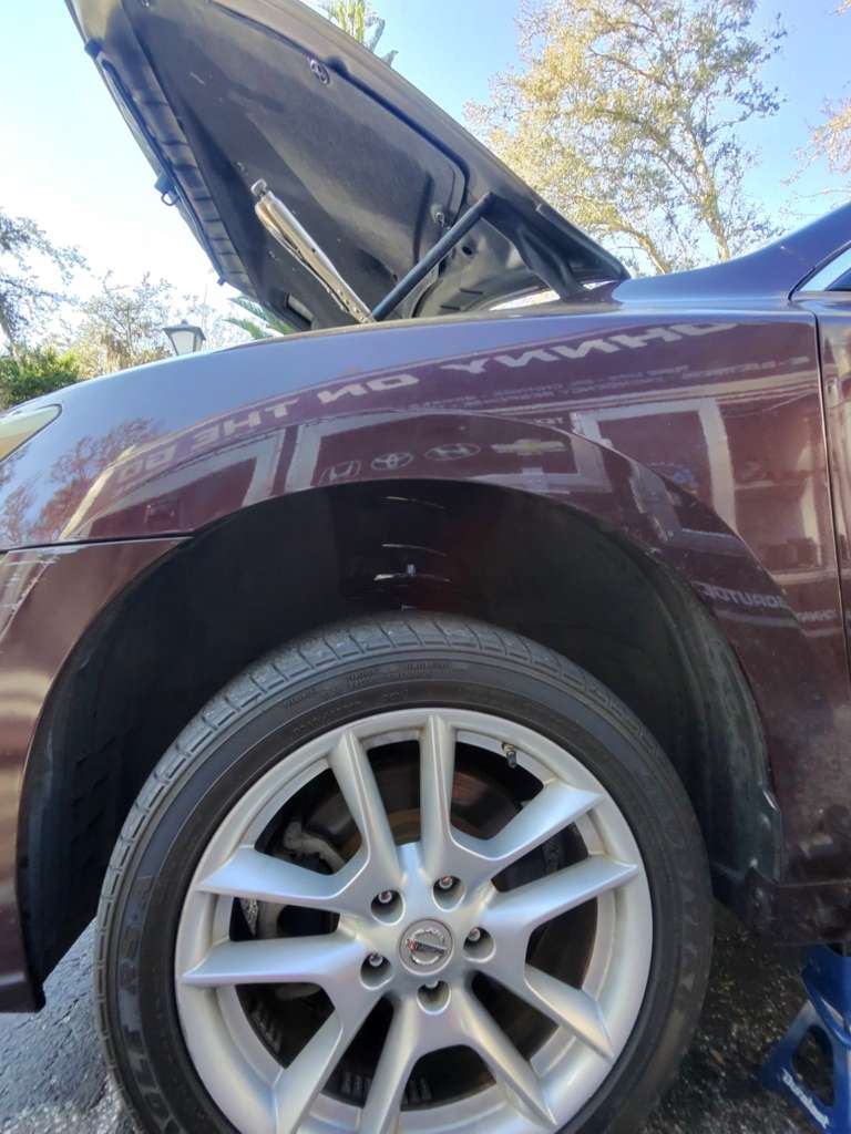 A burgundy vehicle is parked outdoors with its hood open, displaying the engine compartment and a silver alloy wheel, with autumn foliage visible in the background.