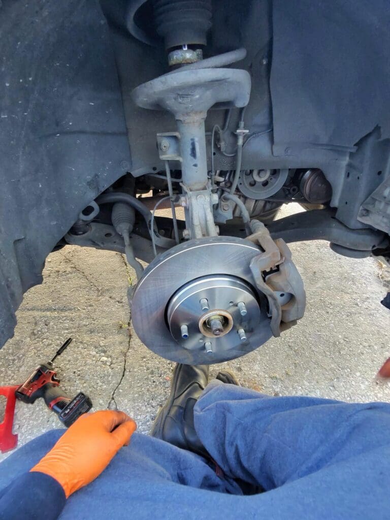 A mechanic performs brake system maintenance on a vehicle, photographed from above showing the brake rotor, caliper, and work in progress.