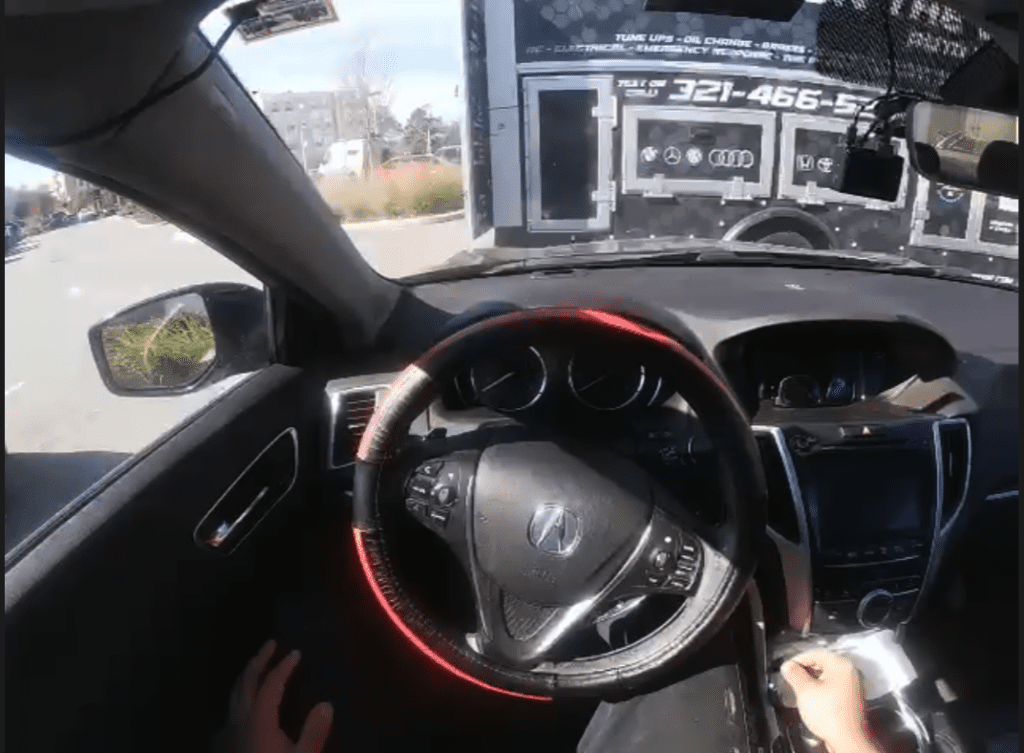 Interior view from the driver's seat of a vehicle showing the steering wheel and dashboard with a road and parked trucks visible through the windshield.