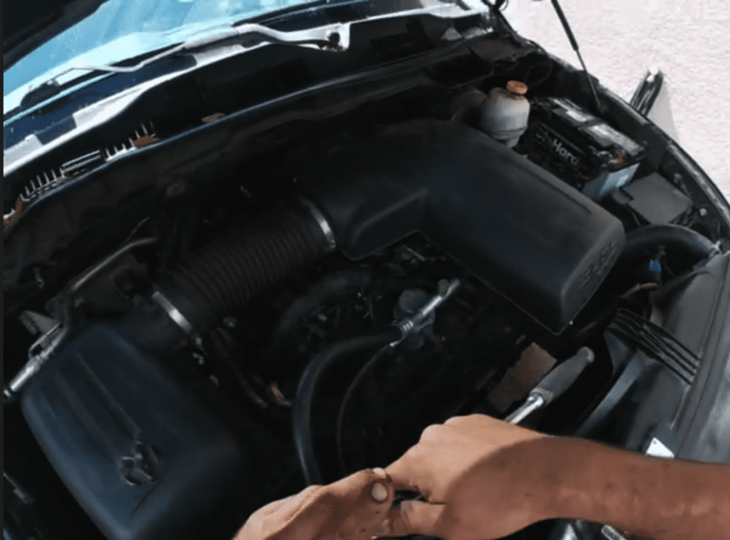 A mechanic or car owner inspects the open engine compartment of a vehicle in daylight, examining the engine block and internal components.