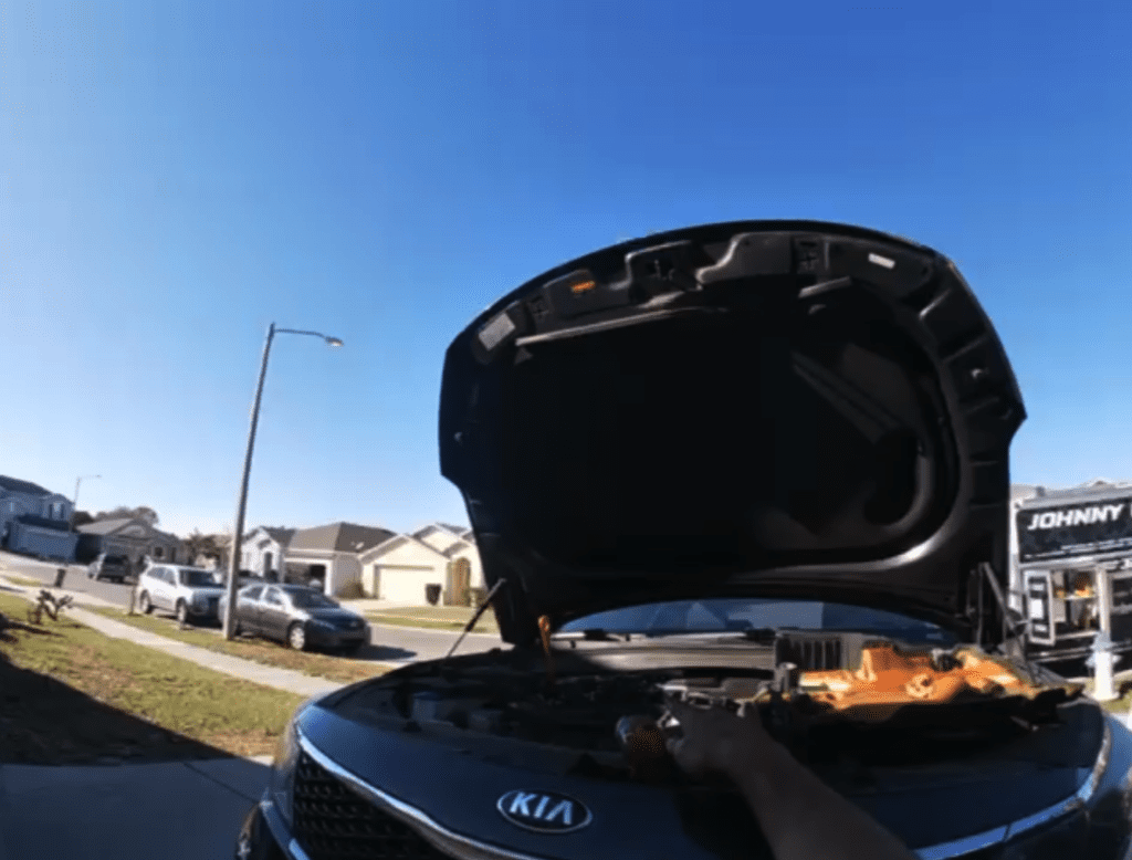 A Kia vehicle's engine compartment is engulfed in flames during daytime in a residential parking area.