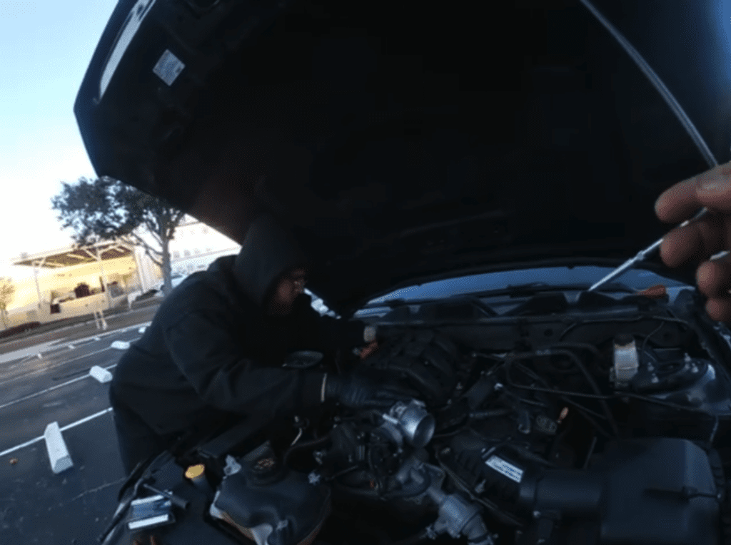 A mechanic works on an open car engine under the hood during daytime, performing maintenance or repairs on the roadside.