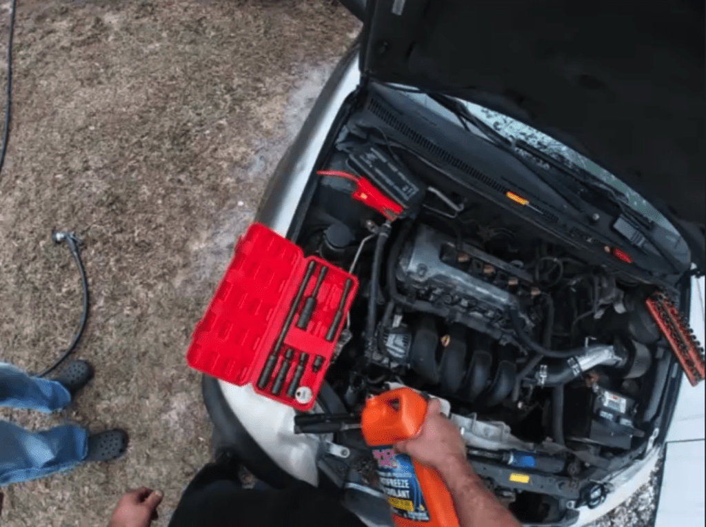 A close-up view of an open car engine bay showing the motor and internal mechanical components with a red hood raised against a concrete workshop floor.