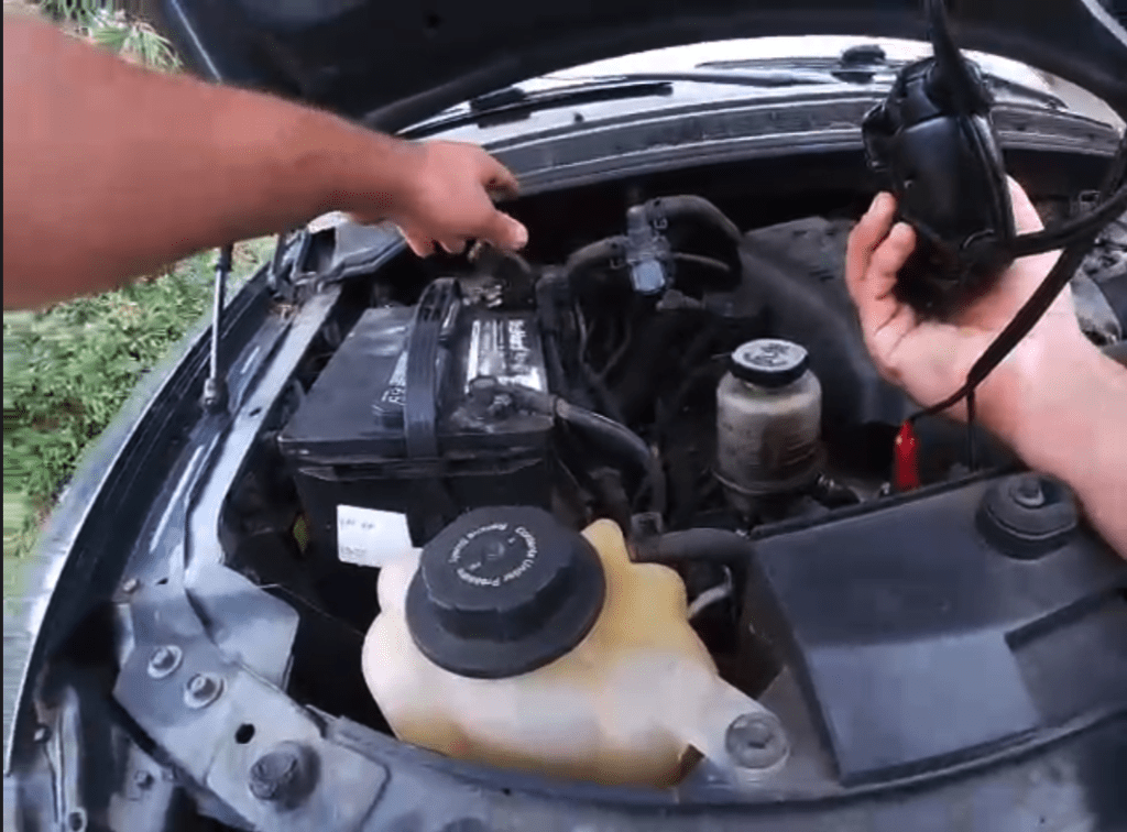 A person performs engine maintenance on a vehicle, checking or refilling the coolant reservoir under the open hood in daylight.