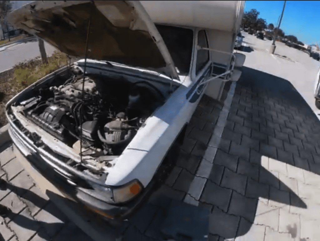 An overhead view of a vintage white car with its hood open, revealing the engine and mechanical components in daylight.