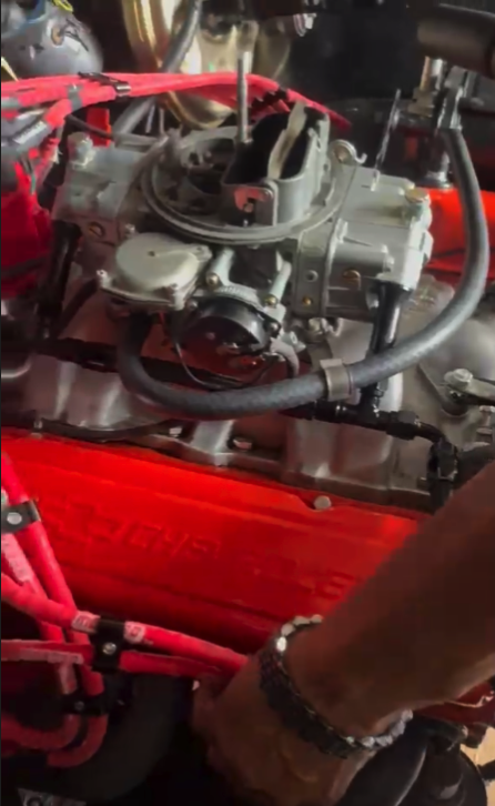 A close-up view of a bright red automotive engine with chrome and metal components visible in detail, likely in a workshop or garage setting.