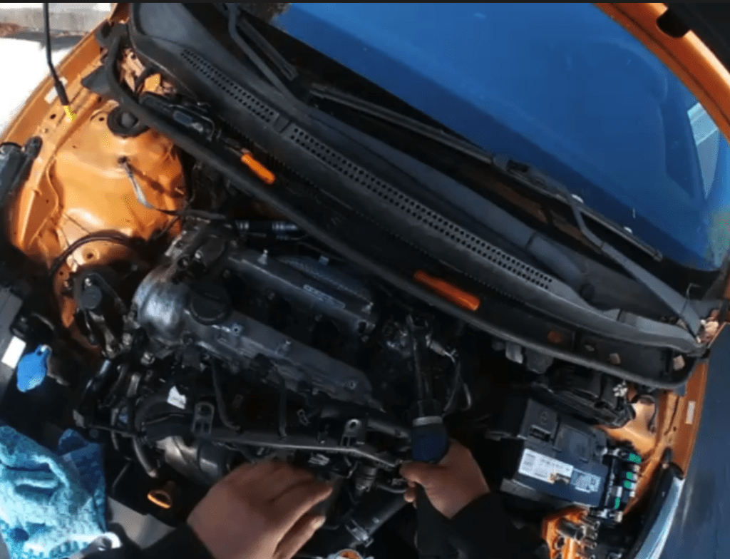 A close-up view of a disassembled car engine with orange bodywork visible above, showing internal mechanical components in a workshop setting.