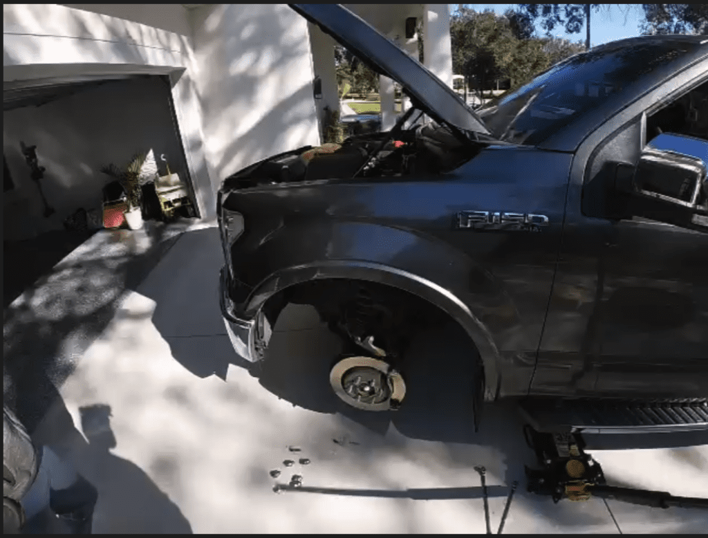 A mechanic works on an open engine hood of a blue pickup truck in an outdoor workshop setting with strong shadow patterns.