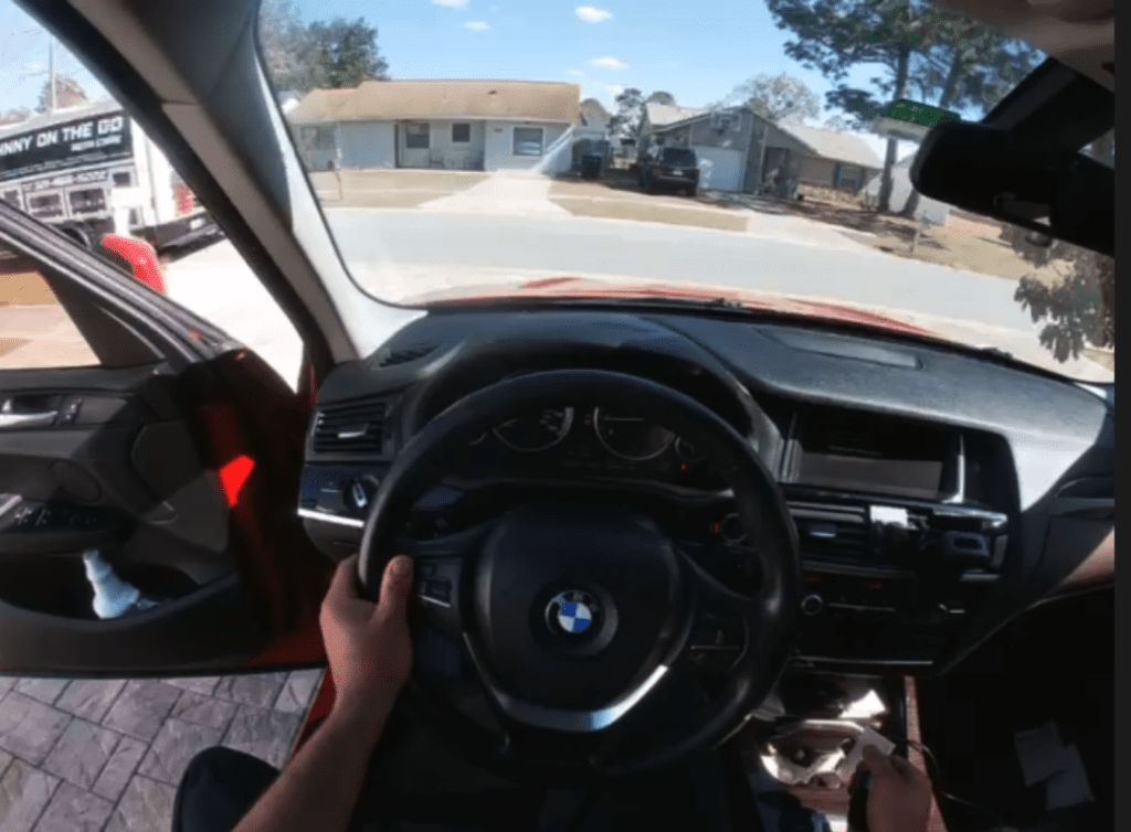 A driver's view from inside a BMW car on a sunny residential street, showing the steering wheel, dashboard, and view through the windshield.
