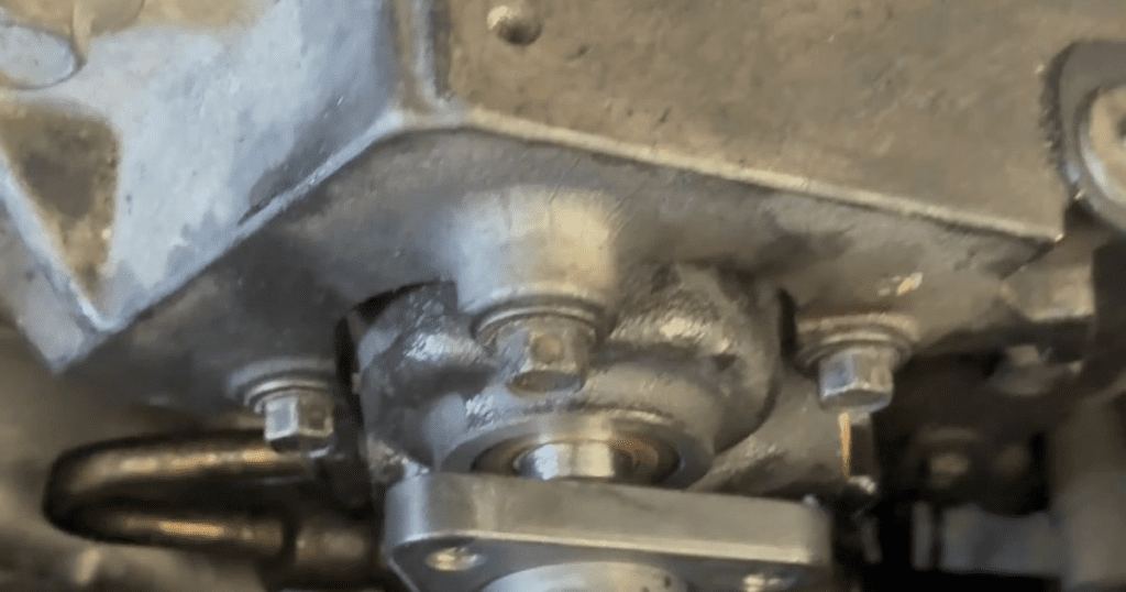 A close-up photograph of a metallic engine component, likely a carburetor or fuel system part, showing detailed mechanical construction with bolts and cast metal surfaces.