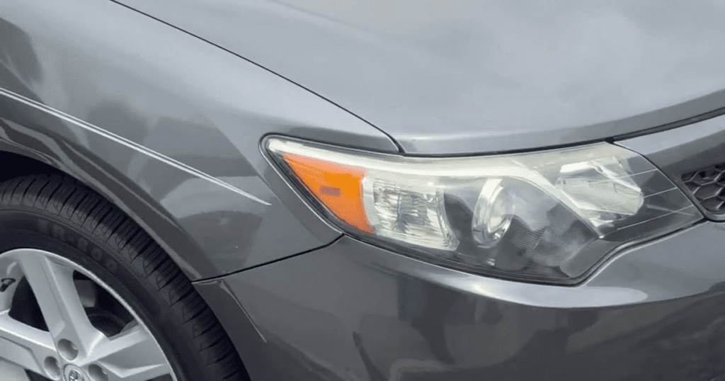 Close-up detail of a gray sedan's front headlight assembly and fender in daylight conditions.