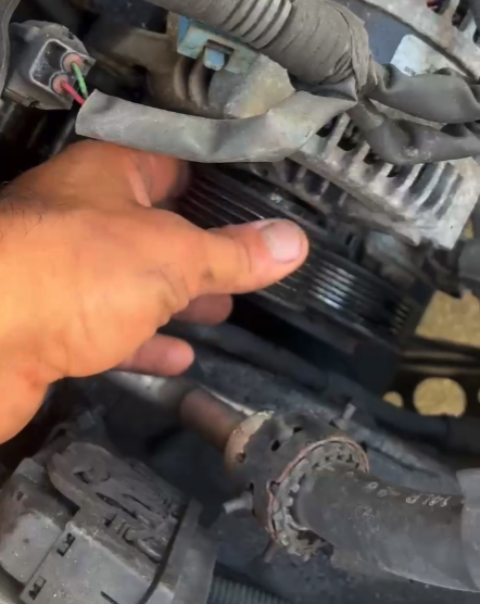 A close-up of a mechanic's hand inspecting or adjusting an engine component within a vehicle engine bay during daytime.