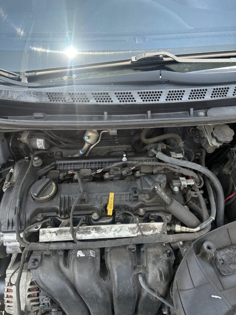 An open car hood reveals the engine compartment in bright daylight, showing mechanical components and metal parts with sunlight glinting off the surface.
