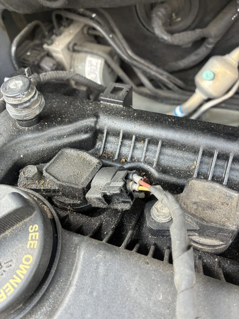 A close-up overhead view of an engine's internal components showing spark plugs, wiring harnesses, hoses, and valve covers in a workshop or garage setting.
