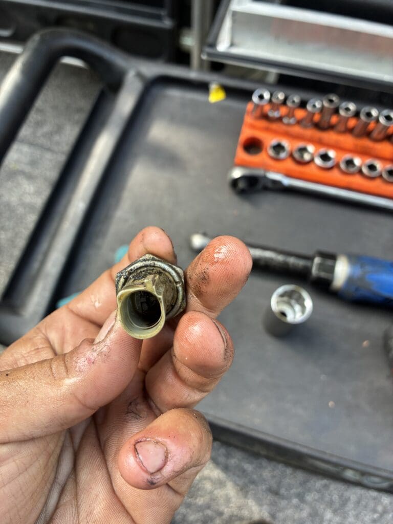 A close-up of a worker's hand holding a dirty brass pipe fitting in a workshop with organized tools visible in the background.