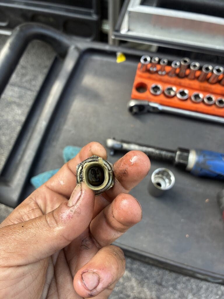 A close-up of a person's hand holding a metallic electrical connector in a workshop setting surrounded by tools and equipment.